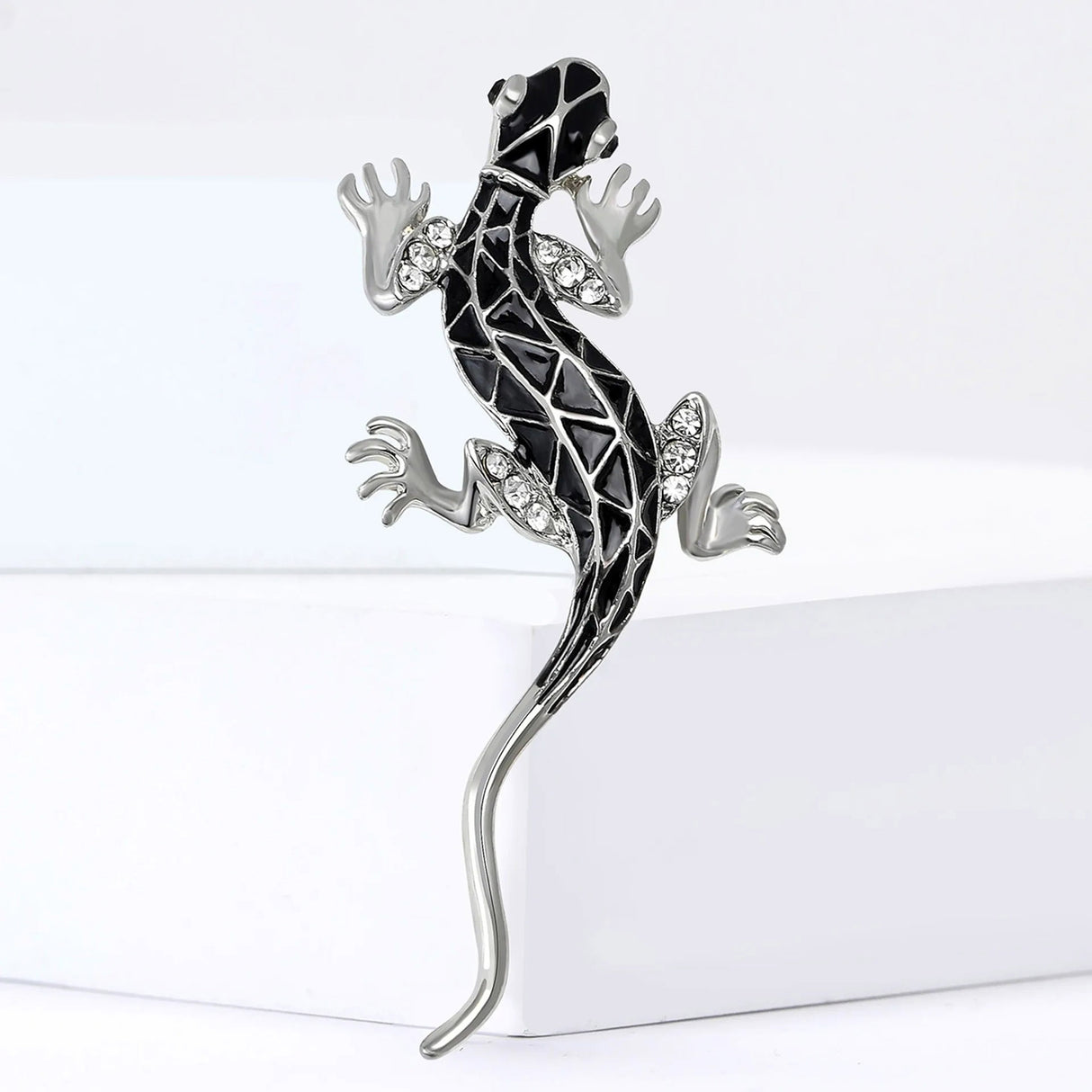 Lizard brooch in silver-tone metal with black enamel geometric design and clear stones, standing on white block.