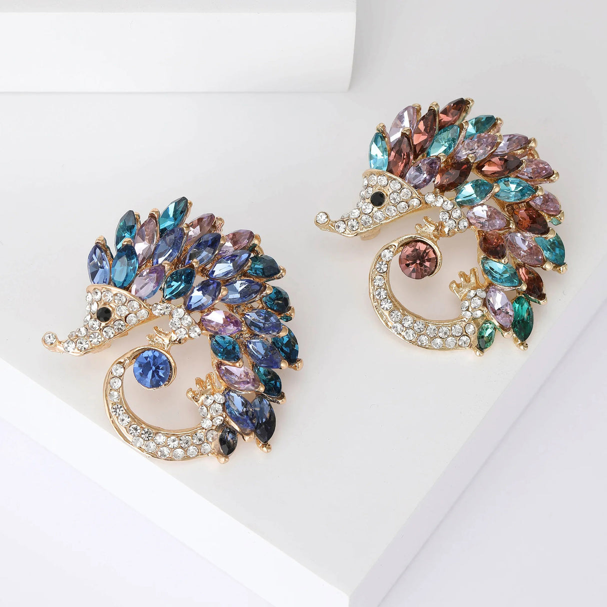 Pair of hedgehog brooches with gold-tone metal, clear small stones, and large multicolor marquise stones, flat lay on white block.