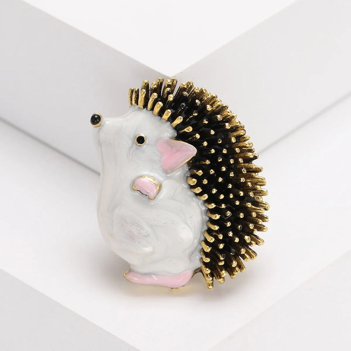 Hedgehog brooch with white body, pink ears and feet, black and gold-tone textured spines, angled view on white.