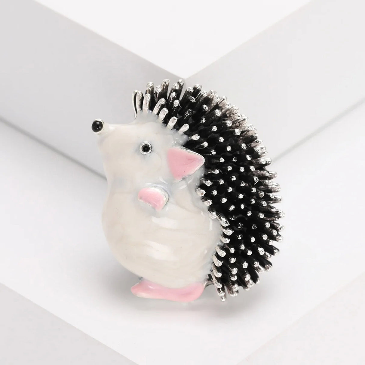Hedgehog brooch with white body, pink ears and feet, black and silver-tone textured spines, angled view on white.