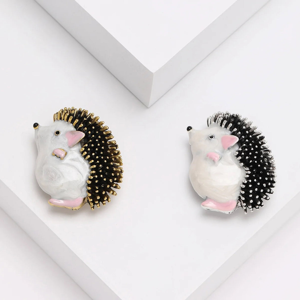 Pair of hedgehog brooches with white bodies and pink accents, one with gold-tone spines and one with silver-tone, angled view.