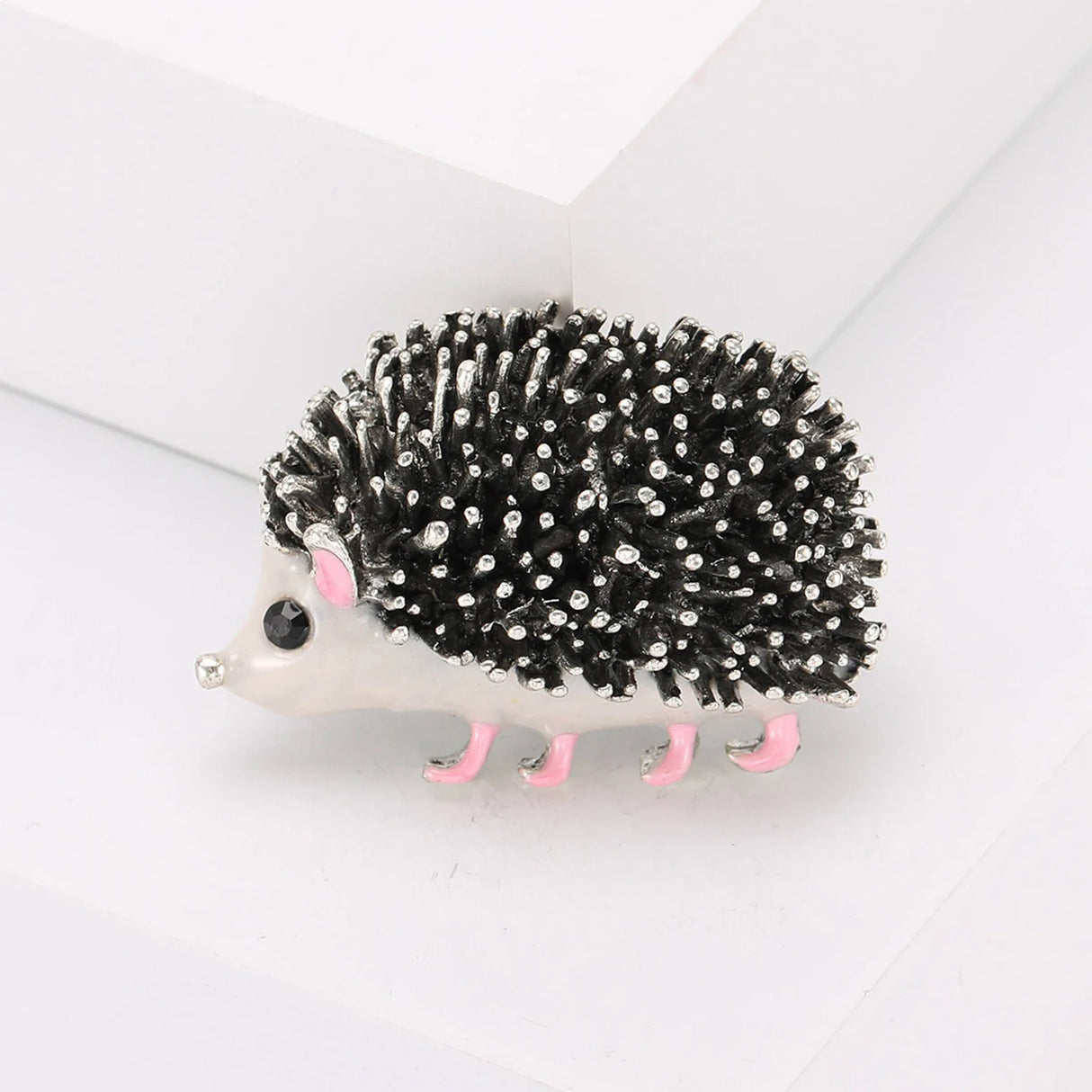 Hedgehog brooch with textured black and silver-tone spines, white body, pink ears and legs. Front view on white block in studio.