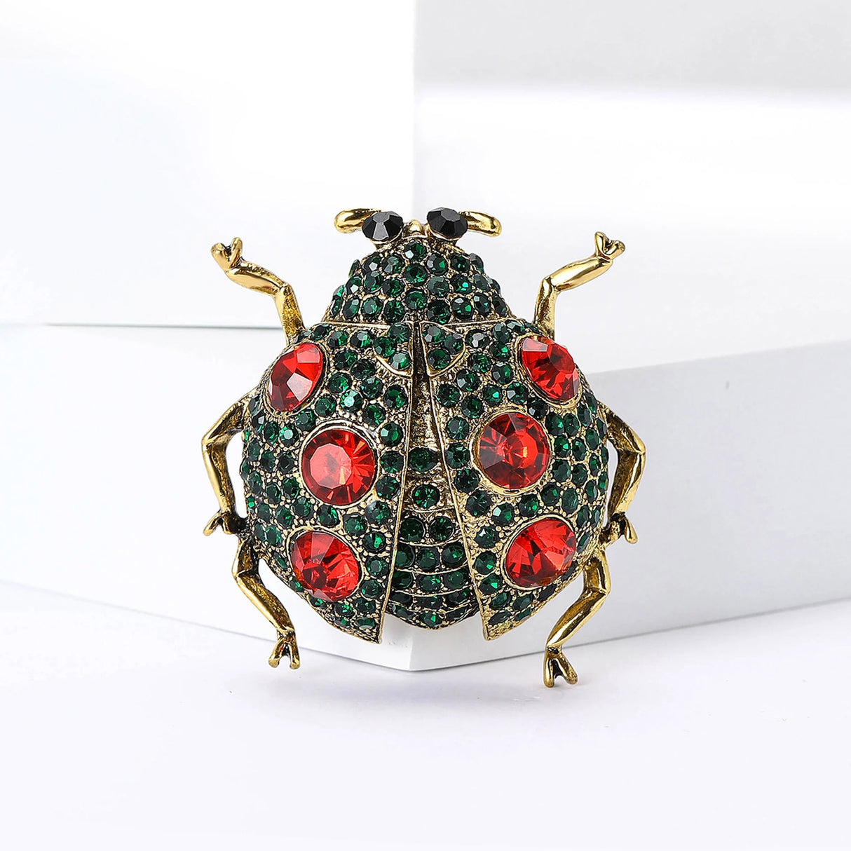 Ladybug brooch with gold-tone metal, green base, red faceted stones, and black eyes, upright pose on white block.