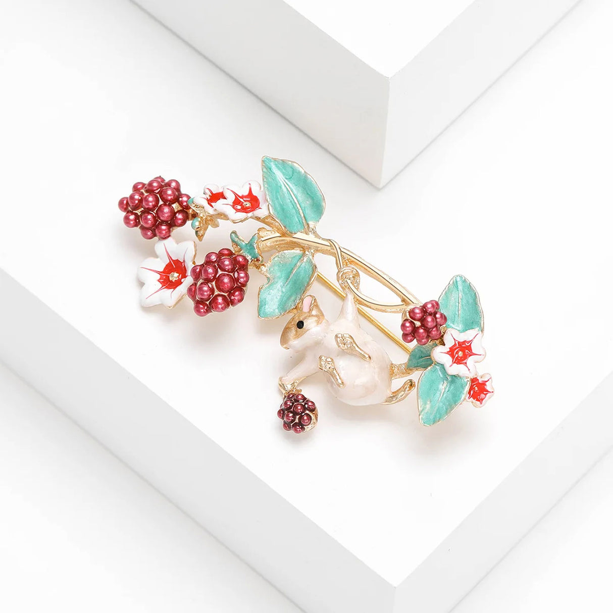 Mouse brooch in gold-tone metal with red berry clusters, green enamel leaves, and star-centered white flowers, angled studio shot.