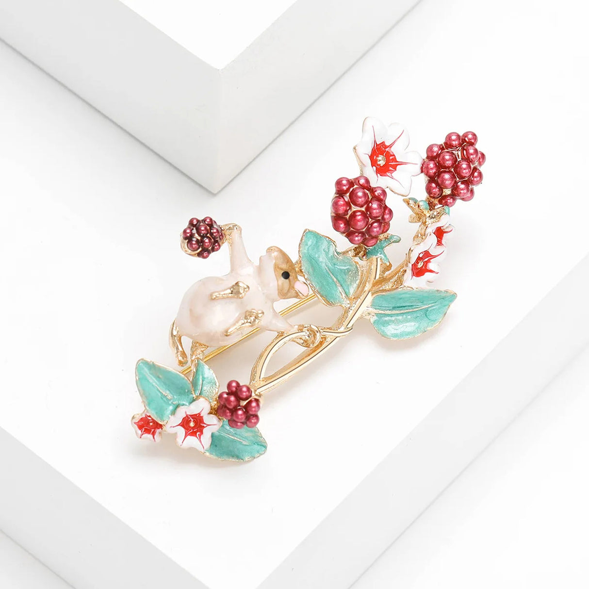 Mouse brooch with white enamel finish, red beaded berries, white flowers with red centers, and green leaves, angled view on white studio platform.