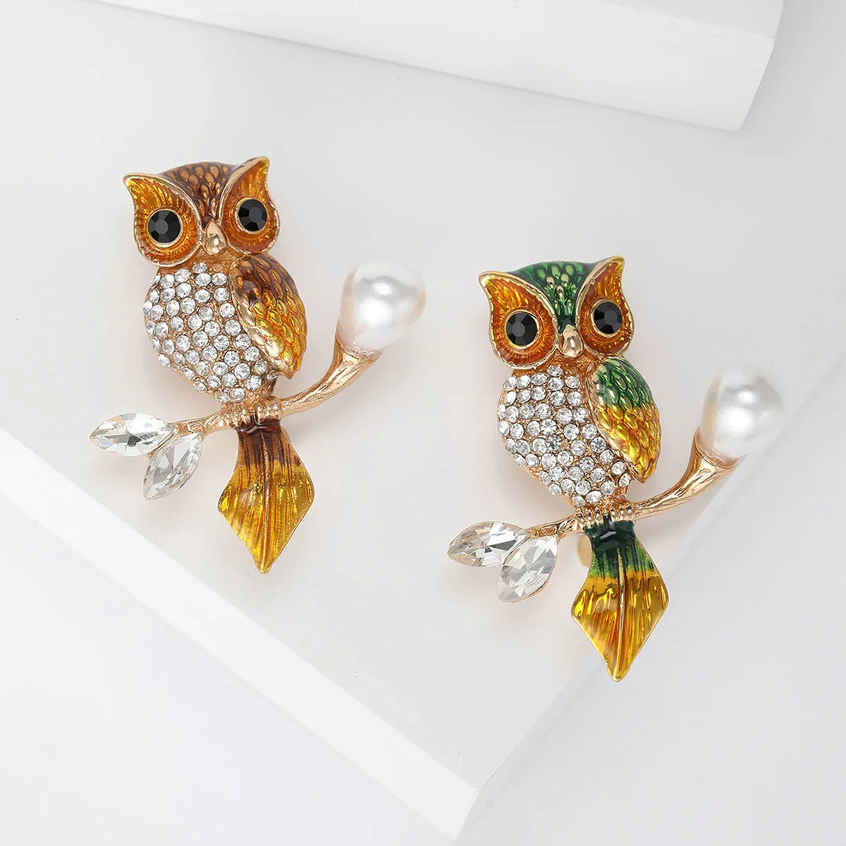 Set of two owl brooches set in gold-tone metal, one in brown and one in green enamel finish with orange accents. Both with clear stones and pearl-like branch detail. Top-down view on white.