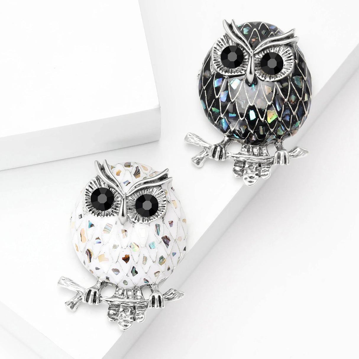 Set of two owl brooches featuring black and white-tone enamel mosaic bodies with reflective accents that resemble abalone shell, top-down view on white blocks.