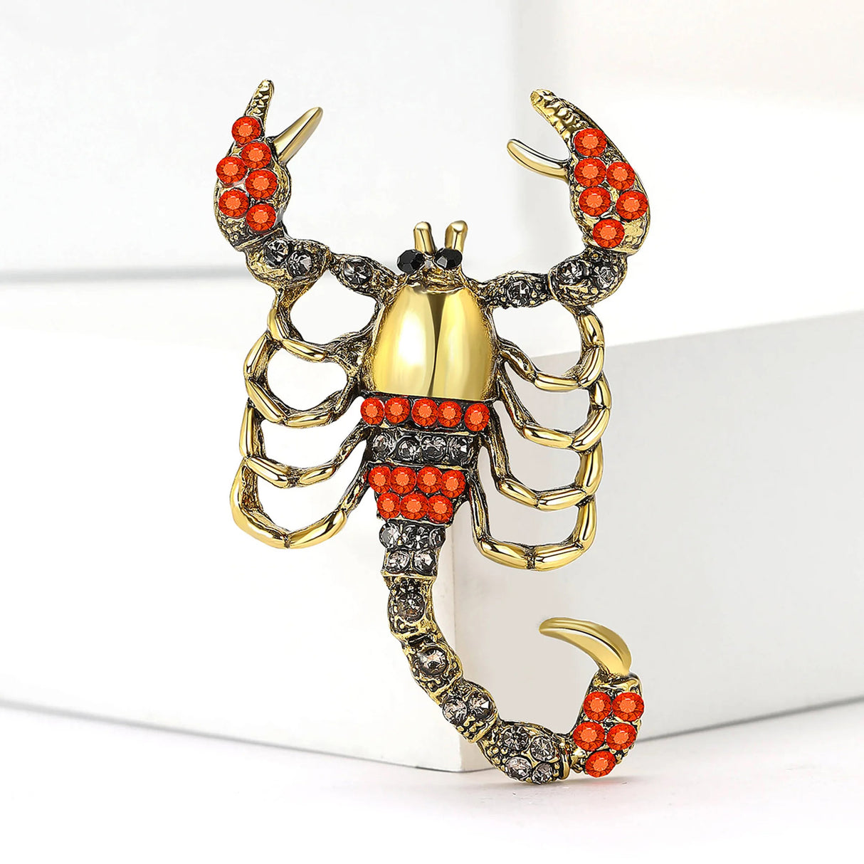 Scorpion brooch with gold-tone metal, orange-red and clear faceted stones, upright display on white block.