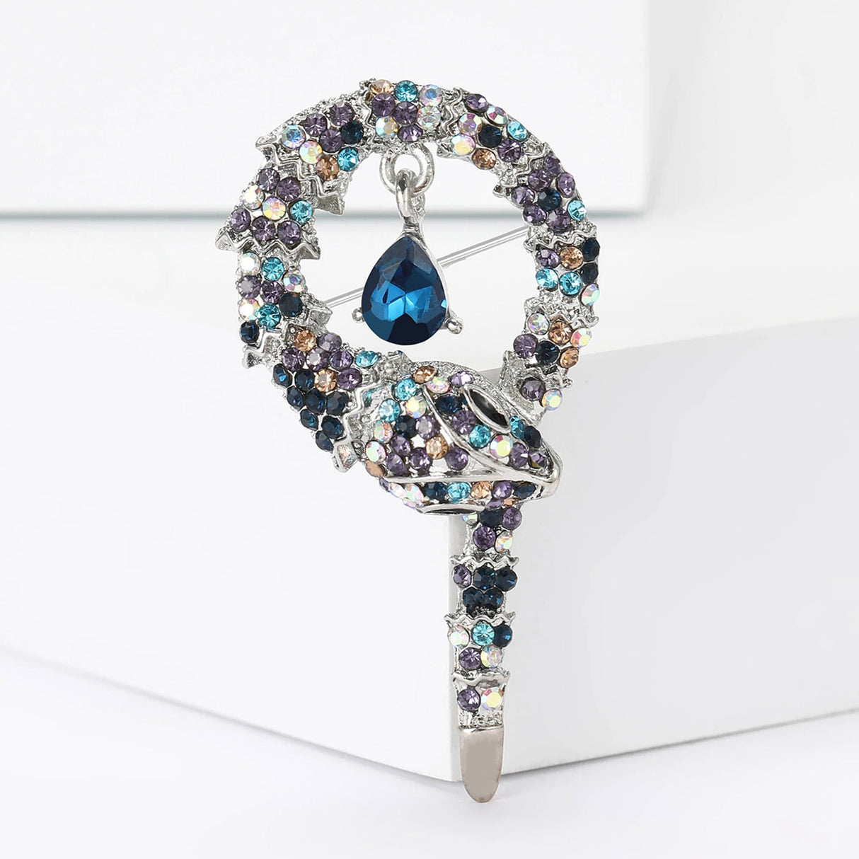 Snake brooch with silver-tone base, multicolor blue-purple-beige iridescent stones, and a hanging blue teardrop stone, upright on white block.