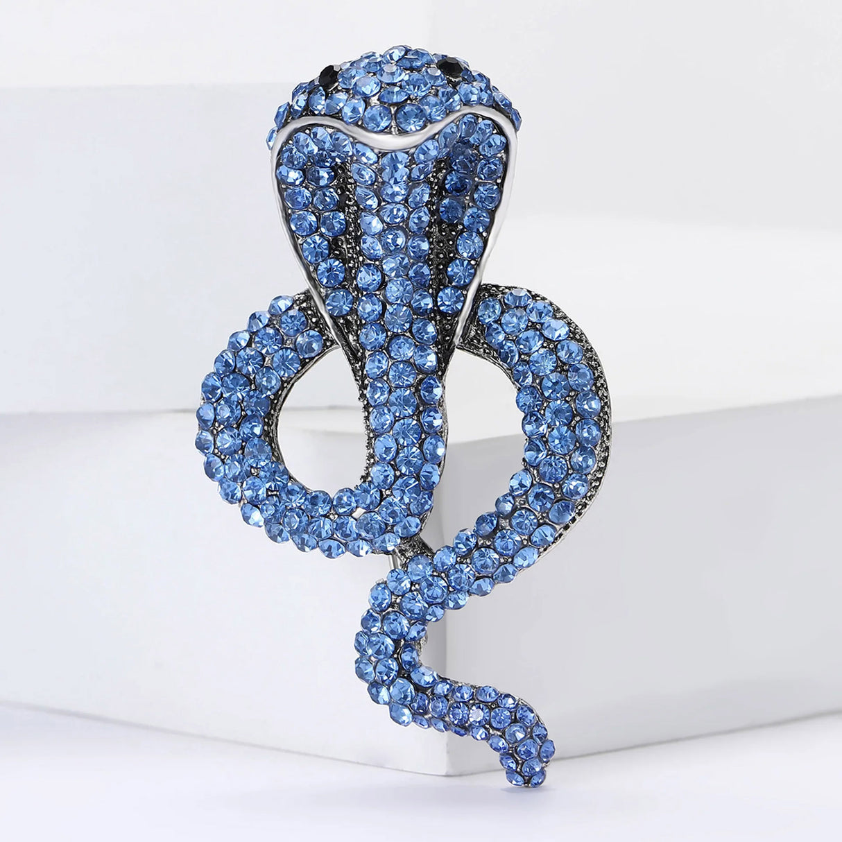 Cobra-shaped snake brooch with blue faceted stones and metallic outline, upright pose on white block background.