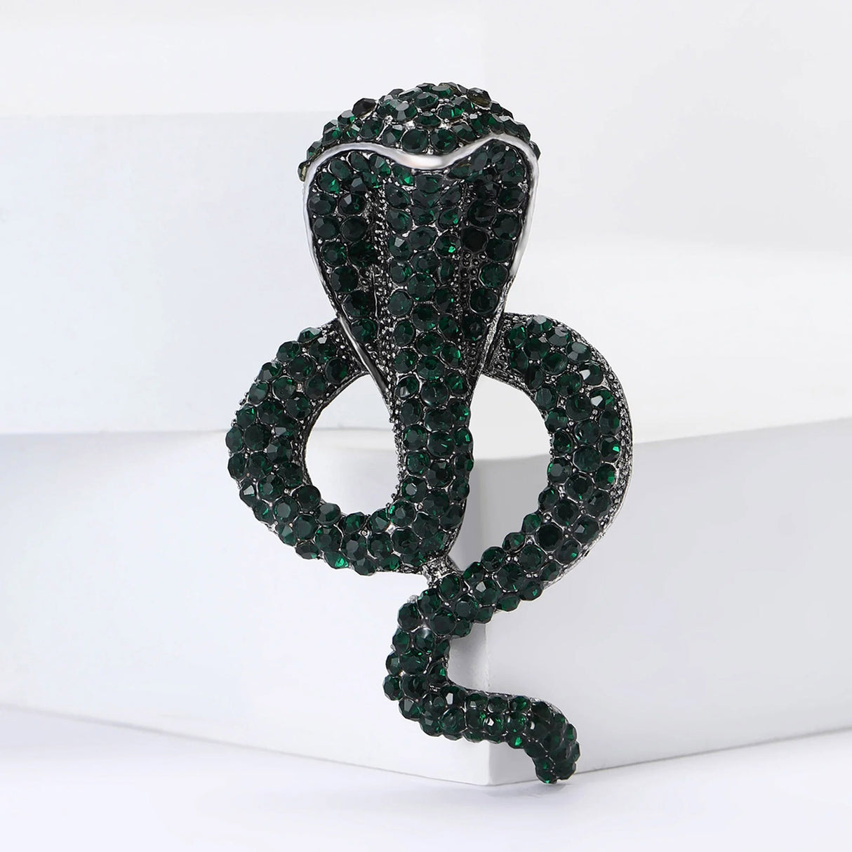 Cobra-shaped snake brooch with dark green faceted stones and silver-tone edging, upright view on white platform.