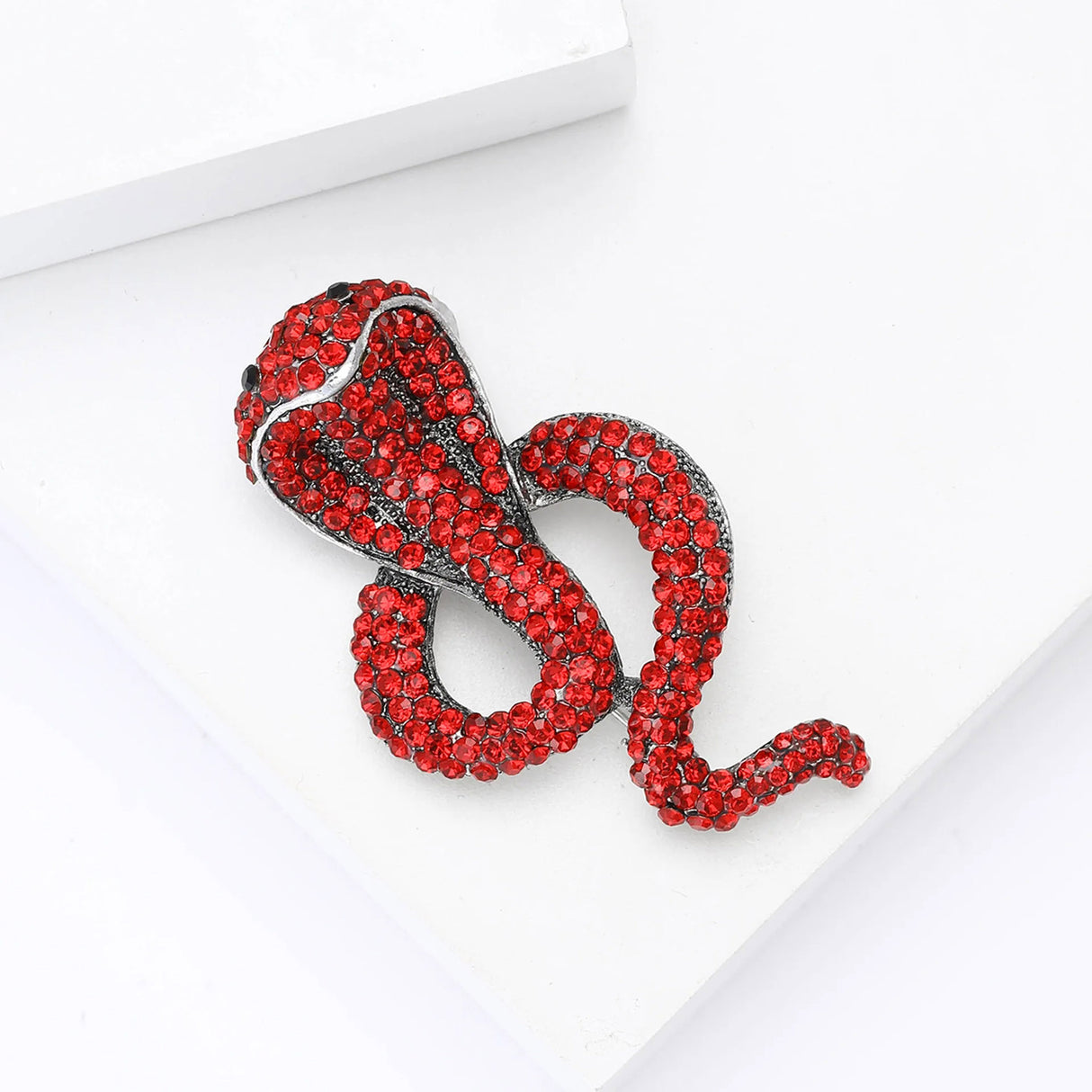 Cobra-shaped snake brooch covered in red pave-set stones with silver-tone edging, angled top-down view on white block.