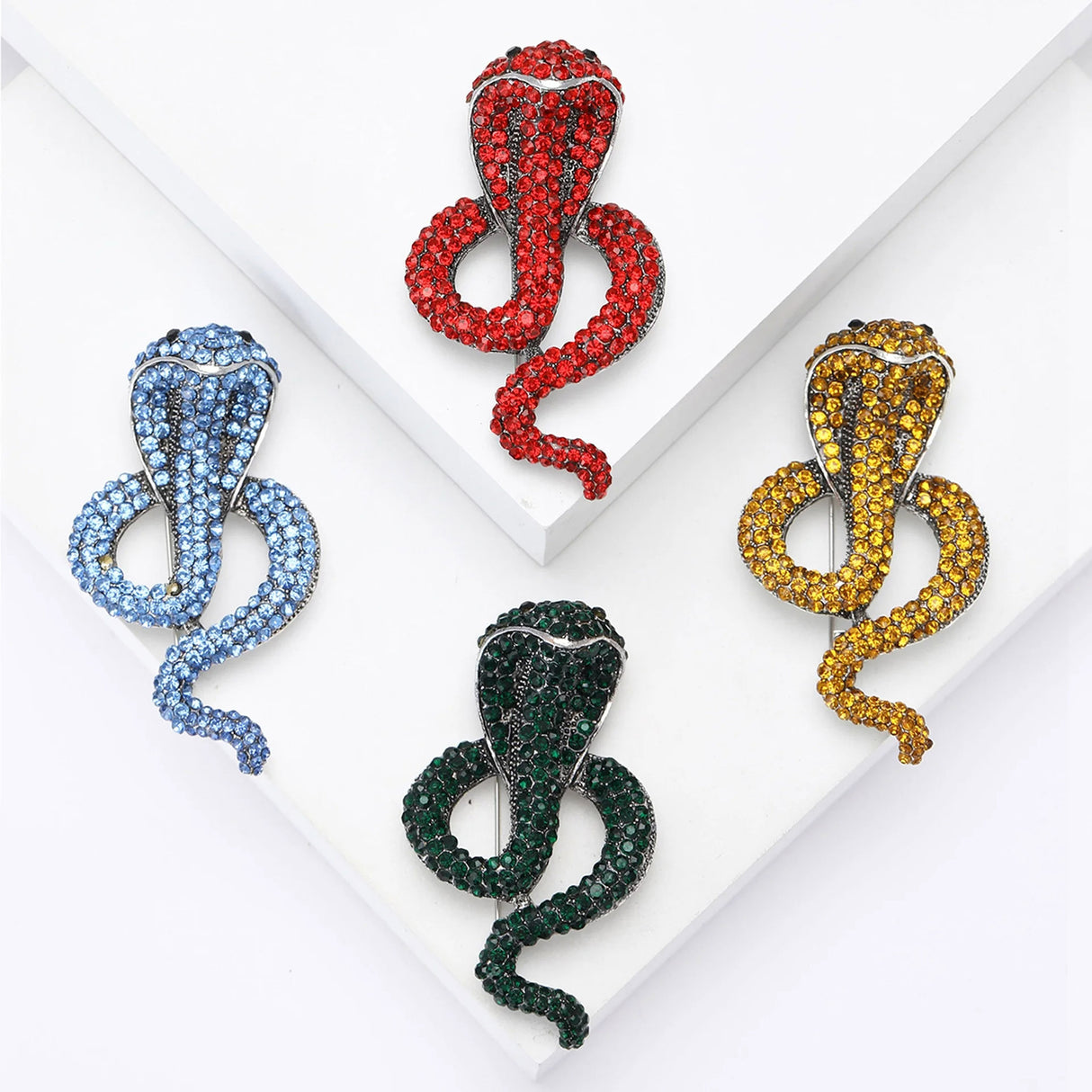Set of cobra snake brooches with blue, red, yellow, and green stones, flat lay on layered white blocks.