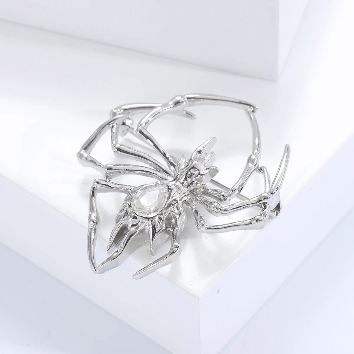 Silver-tone metal spider brooch with clear teardrop-shaped faceted stone as body, angled studio pose on white platform.