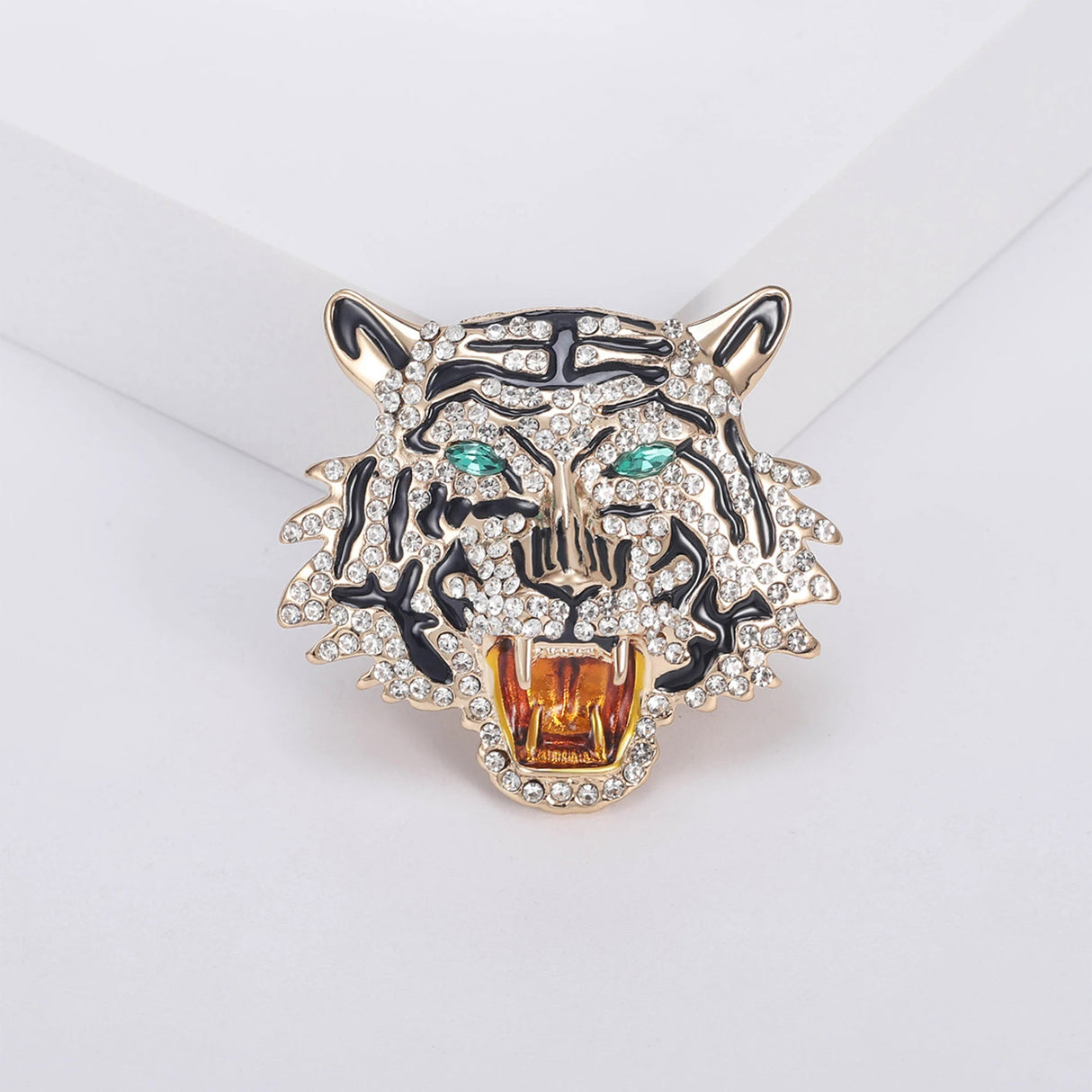 Roaring tiger head brooch with gold-tone base, clear faceted stones, black enamel stripes, and green-tone eyes, displayed on white platform.