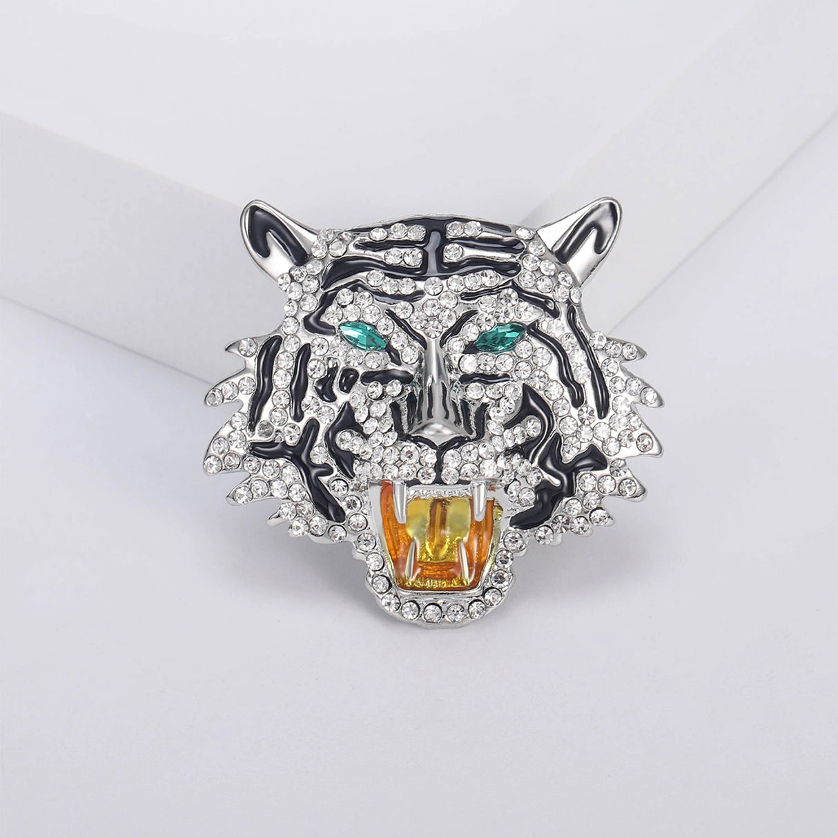 Roaring tiger head brooch with silver-tone base, clear faceted stones, black enamel details, and green-tone eyes, displayed on white platform.