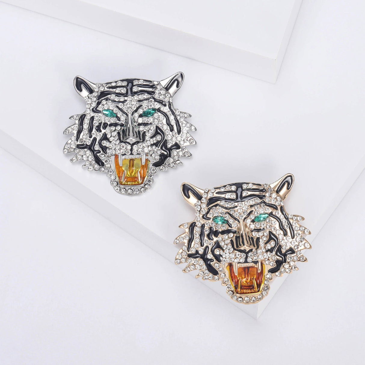 Two roaring tiger head brooches in gold-tone and silver-tone finishes with clear stones, black enamel, and green-tone eyes, flat lay on white blocks.
