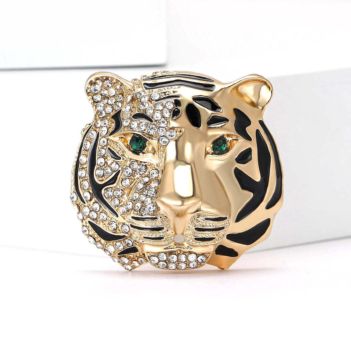 Tiger head brooch with gold-tone finish, half encrusted with clear stones and black enamel, green-tone eyes, standing on white surface.