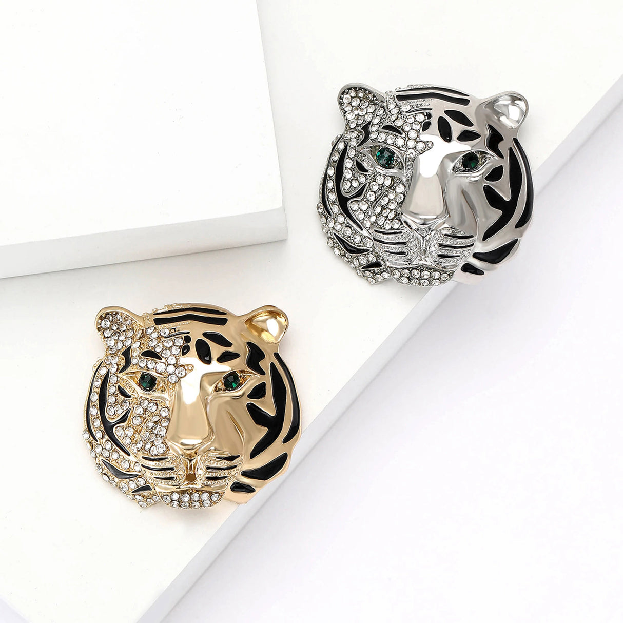 Pair of tiger head brooches in gold-tone and silver-tone finishes, half-studded with clear stones, black enamel detailing, flat lay on white blocks.