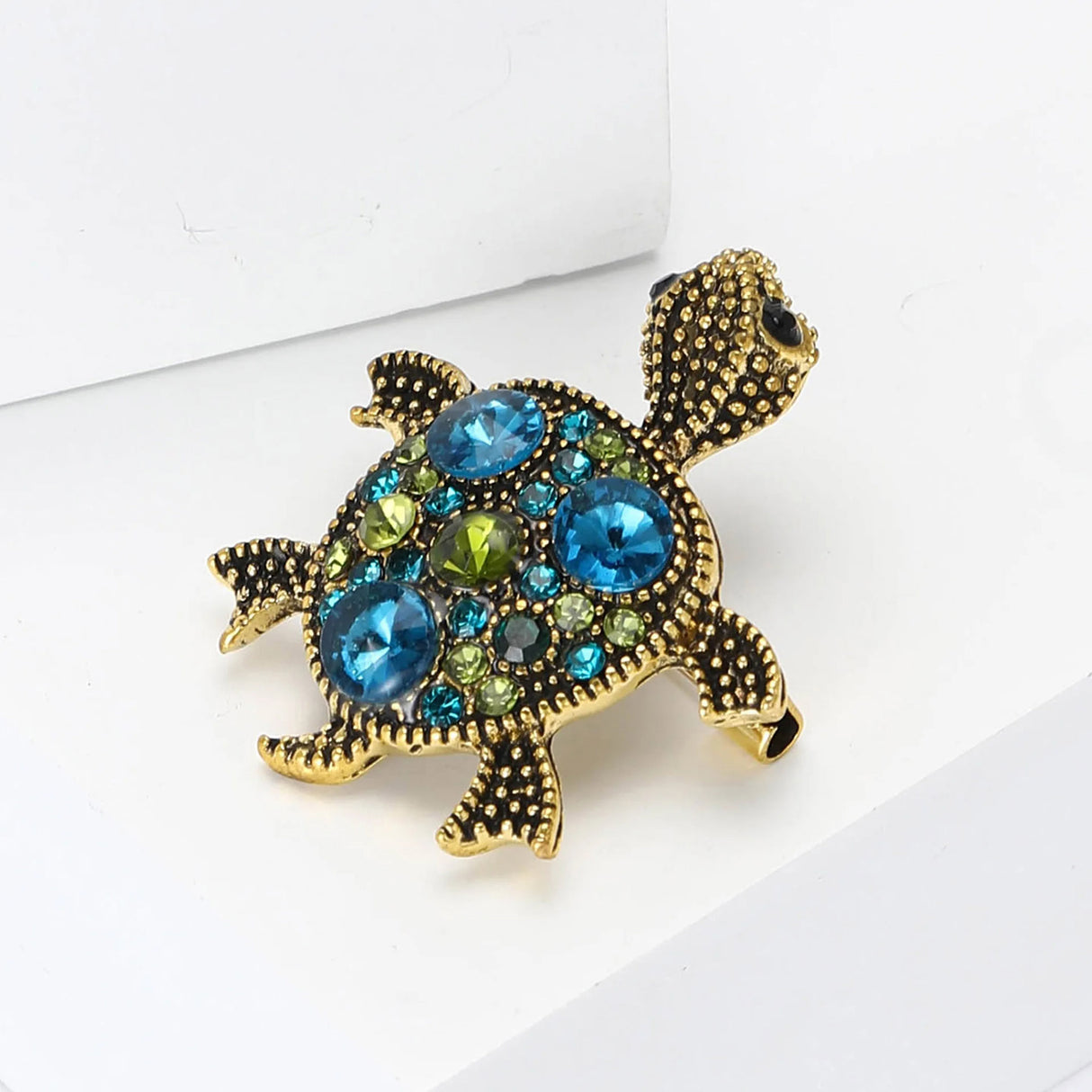 Turtle brooch with gold-tone textured surface and blue-green decorative stones, angled view on white surface.