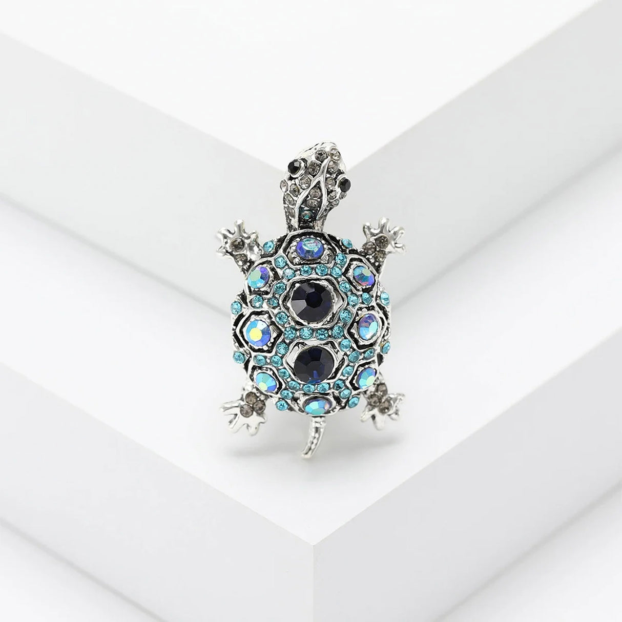 Tortoise brooch with silver-tone metal frame, dark blue faceted center stones, and light blue accents, standing on white stepped platform.