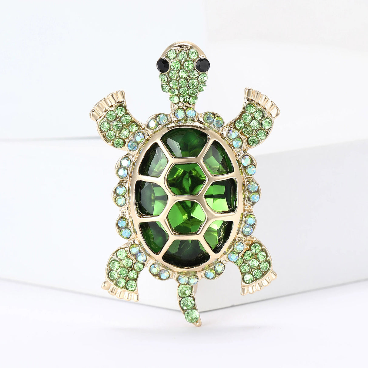 Turtle brooch featuring deep green center stone, pale green round accents, and gold-tone metal, standing on white surface.