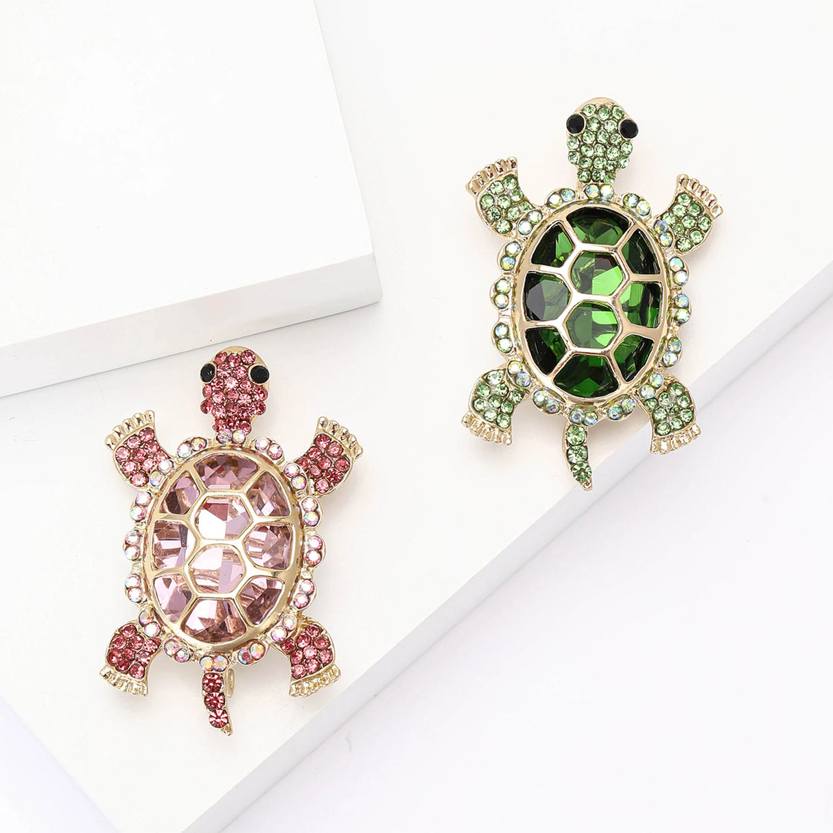 Pair of tortoise brooches with pink and green faceted center stones and small round accents, set in gold-tone metal. Flat lay on white blocks in studio.