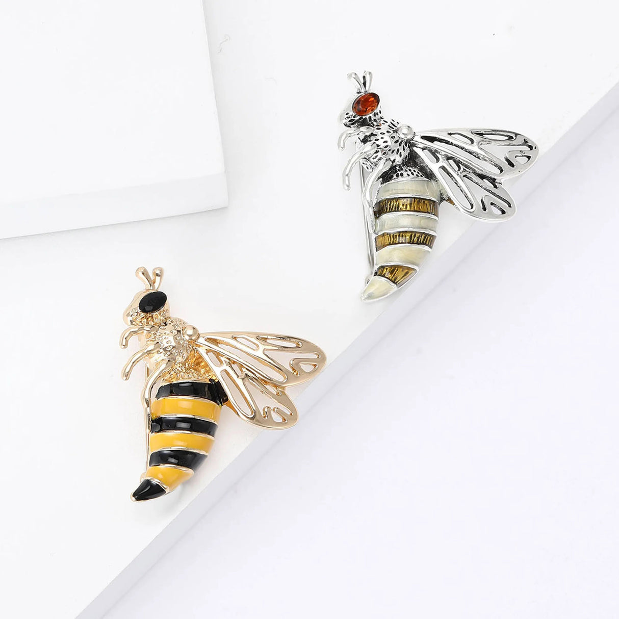 Set of two wasp brooches in gold-tone and silver-tone finishes with striped enamel bodies and openwork wings, top-down view on white surface.