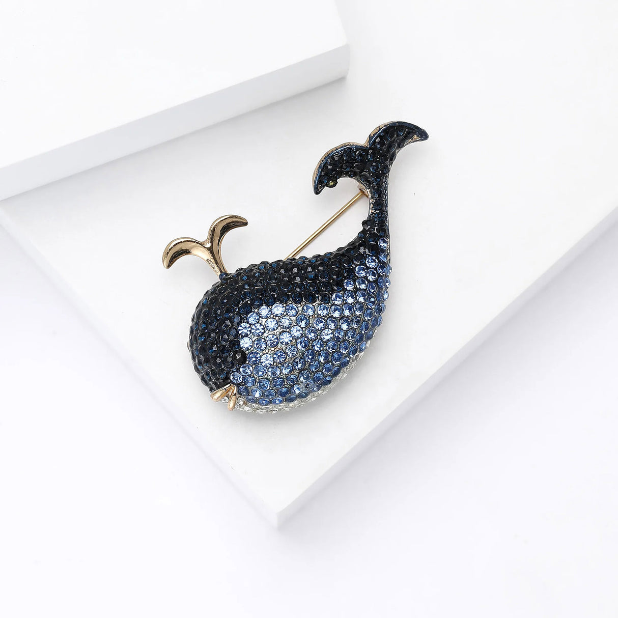 Whale brooch with blue faceted stones and gold-tone water spout accent, angled top-down view on white studio surface.