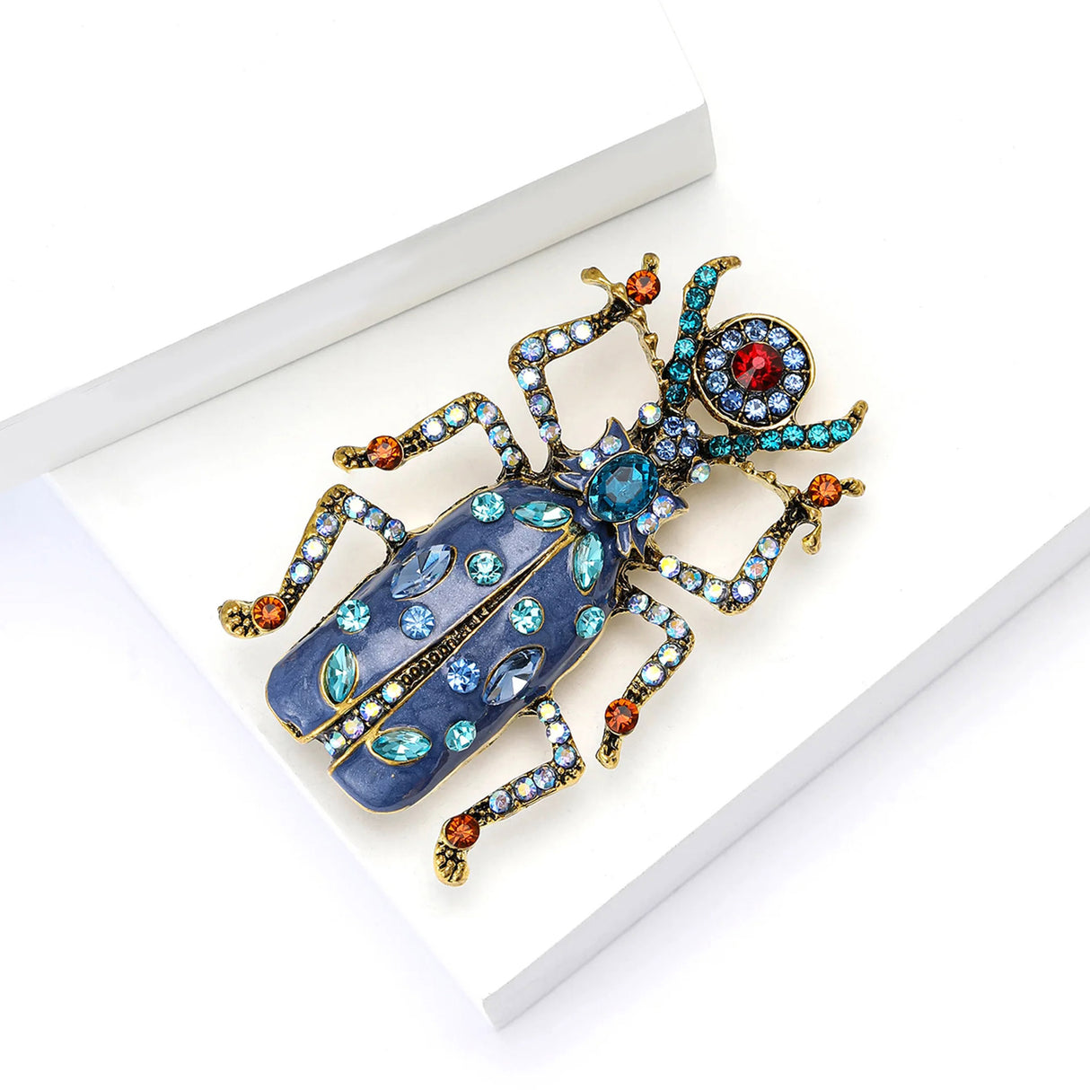 Beetle brooch with blue body, gold-tone legs, and blue and red faceted stones on wings and limbs, angled view on white.
