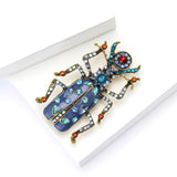 Beetle brooch with blue body, gold-tone legs, and blue and red faceted stones on wings and limbs, angled view on white.
