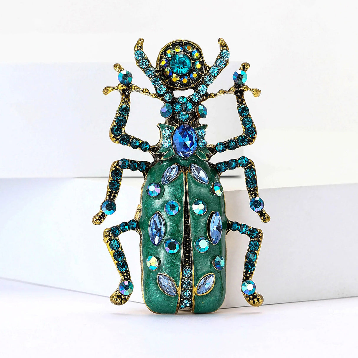 Beetle brooch with blue-green enamel body, gold-tone limbs, and various blue-toned stones across the wings and head, upright front view.