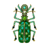 Beetle brooch with green enamel wings, light green and teal stones, and gold-tone metal finish, cutout on white.