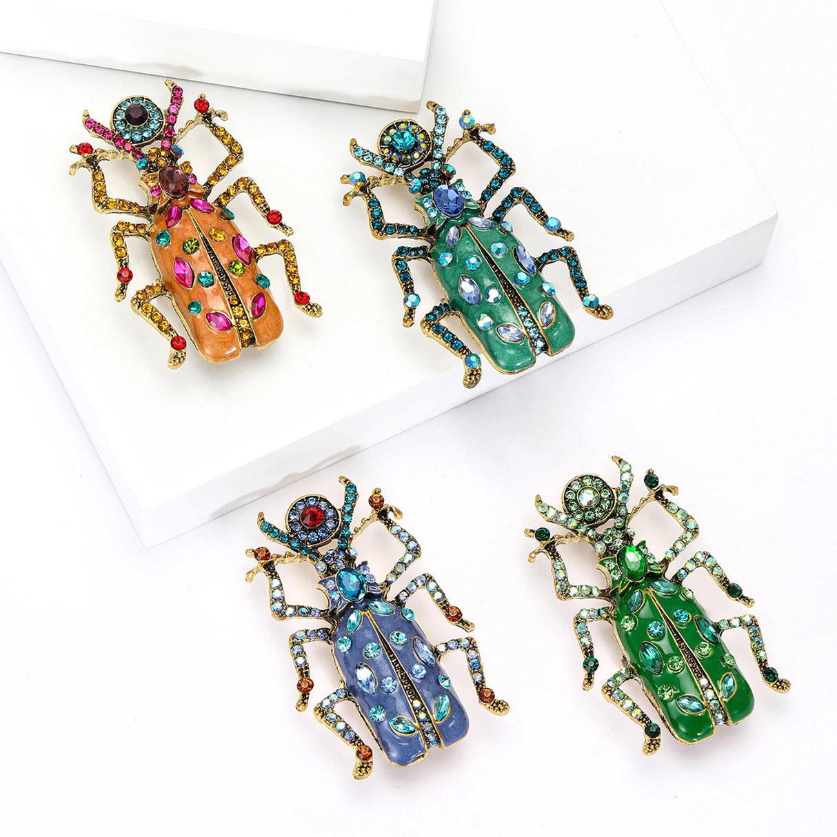 Set of four beetle brooches in assorted body colors with gold-tone limbs and multicolor decorative stones, flat lay on white.