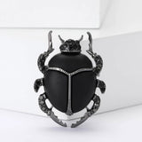 Beetle brooch with matte black body, gray metallic outline, and black faceted stones, upright pose on white block.