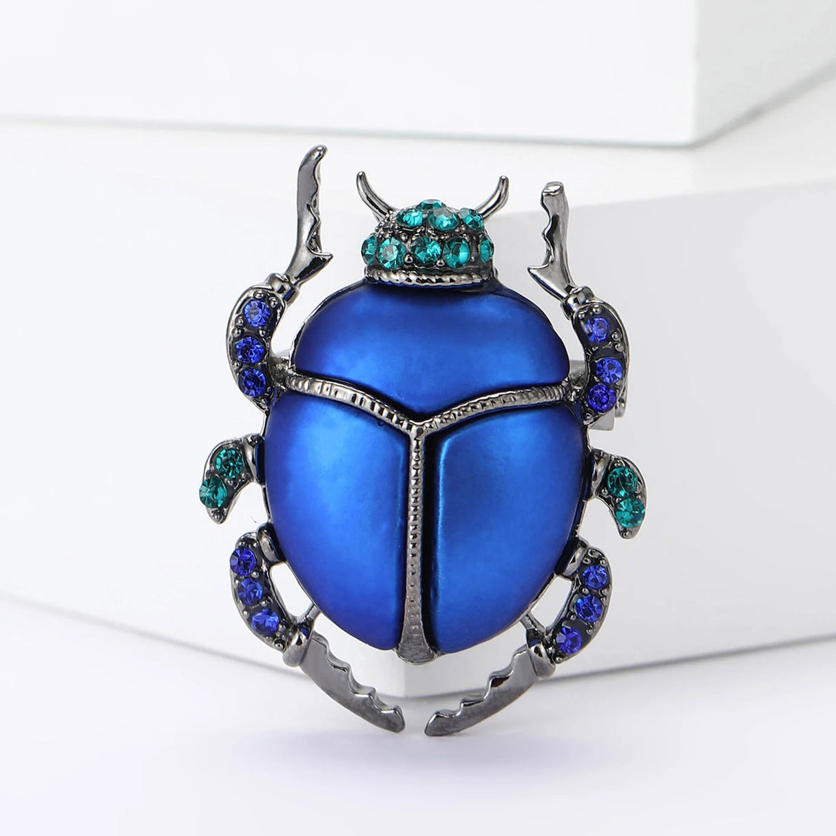 Beetle brooch with metallic blue body, dark metal edges, and blue and green faceted stones, upright view on white.
