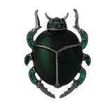 Beetle brooch with green body, dark metallic legs, and green faceted stones, cutout on white background.