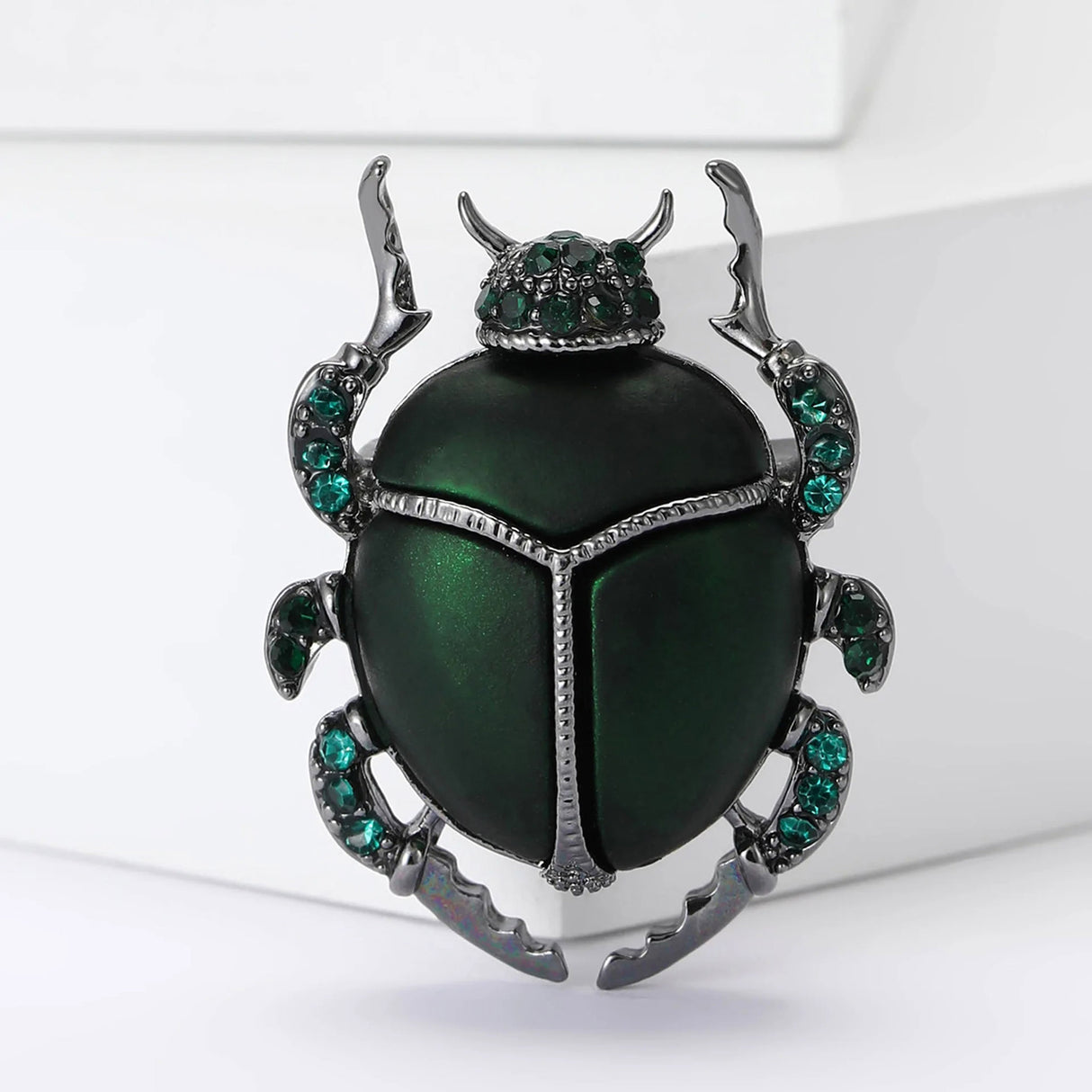 Beetle brooch with green body, dark metallic frame, and green faceted stones, upright view on white surface.