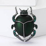 Beetle brooch with green body, dark metallic frame, and green faceted stones, upright view on white surface.