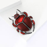 Beetle brooch with red enamel body, gray metal base, and red stone accents, angled bird's eye view.