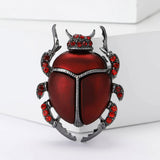 Beetle brooch with metallic red body, dark silver outline, and small round red stones, upright studio pose on white.