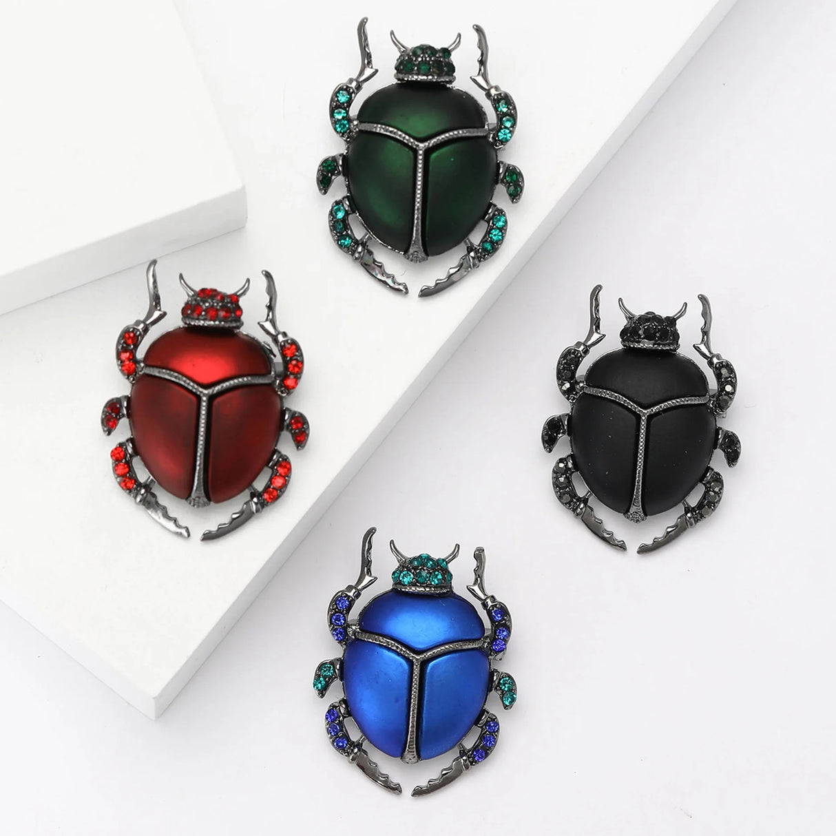 Set of four beetle brooches in black, red, green, and blue metallic enamel, with gray metal legs accented with small stones in matching colors. Flat lay on white background.