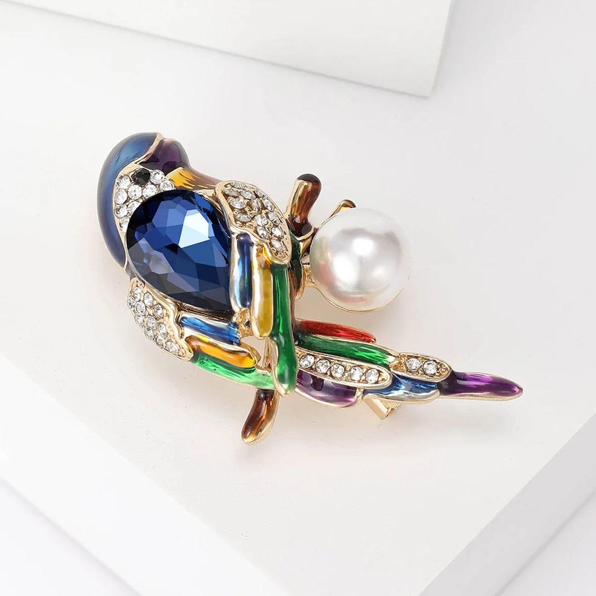 Parrot brooch with blue faceted stone, rainbow enamel details, clear pavé stones, and round white bead, angled side view on white surface.