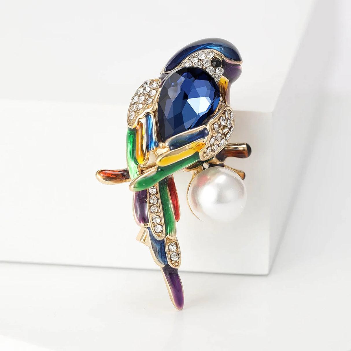 Parrot brooch with blue faceted centerpiece, multicolor enamel accents, clear stones, and round white imitation pearl, upright pose on white block.