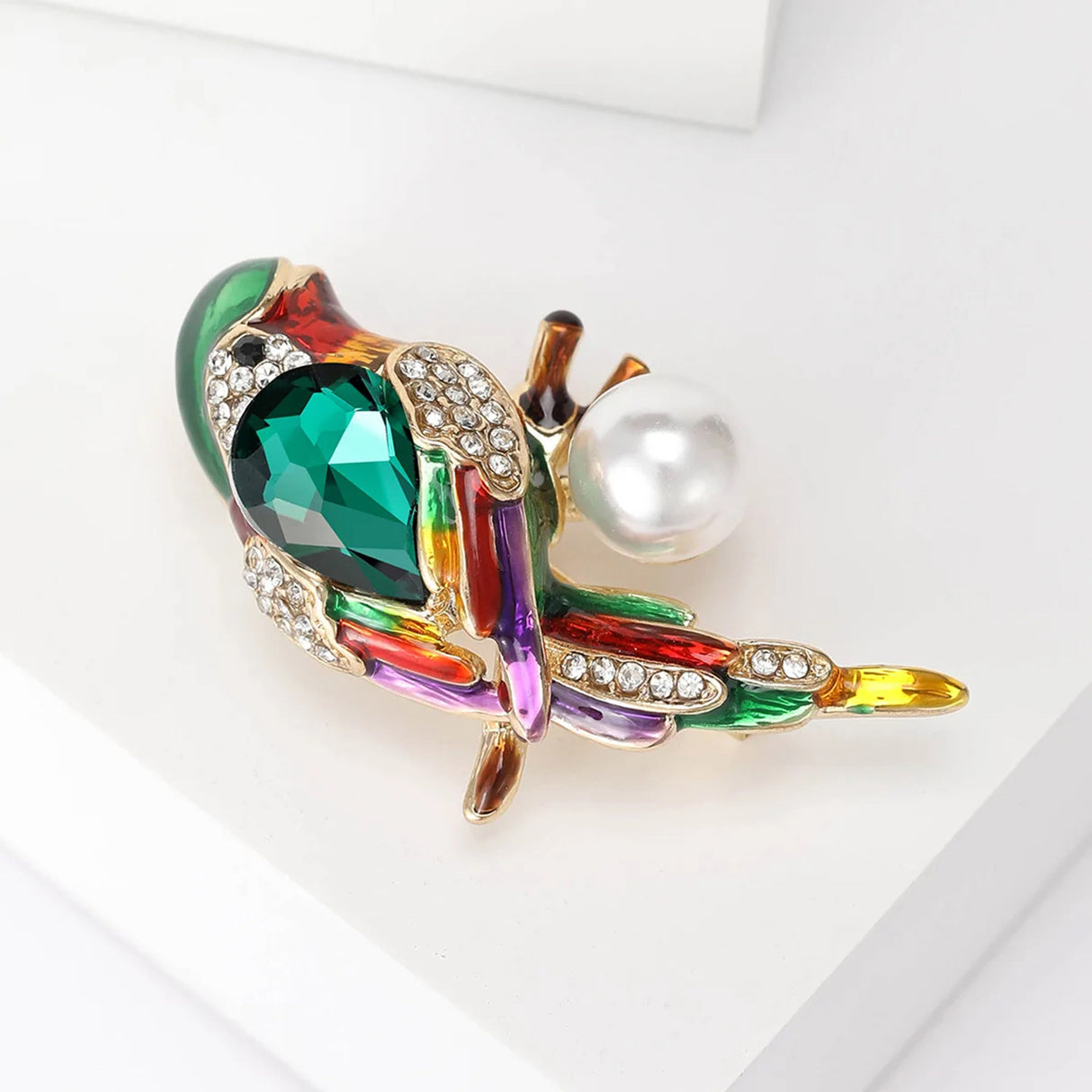 Parrot brooch with green faceted stone, multicolor enamel details, clear pavé stones, and round white bead, angled side view on white block.