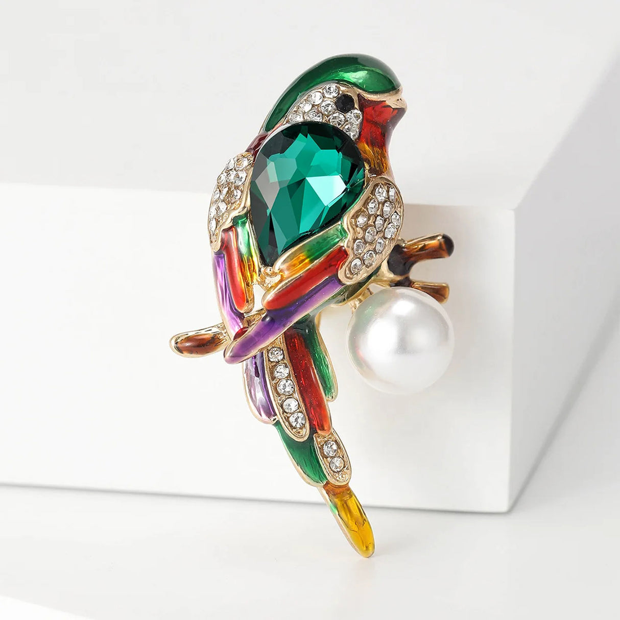 Parrot brooch with green faceted centerpiece stone, rainbow enamel finish, clear pavé stones, and round white faux pearl, upright view on white surface.