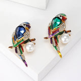 Pair of parrot brooches with blue and green faceted stones, multicolor enamel details, clear pavé accents, and white round beads, flat lay on white.