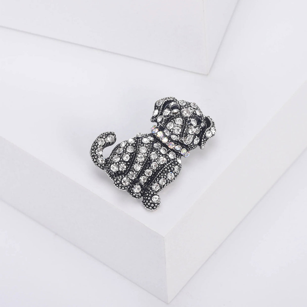 Dog brooch with clear faceted stones, black eyes, and iridescent collar on antique-style silver-tone metal, angled view on white block.
