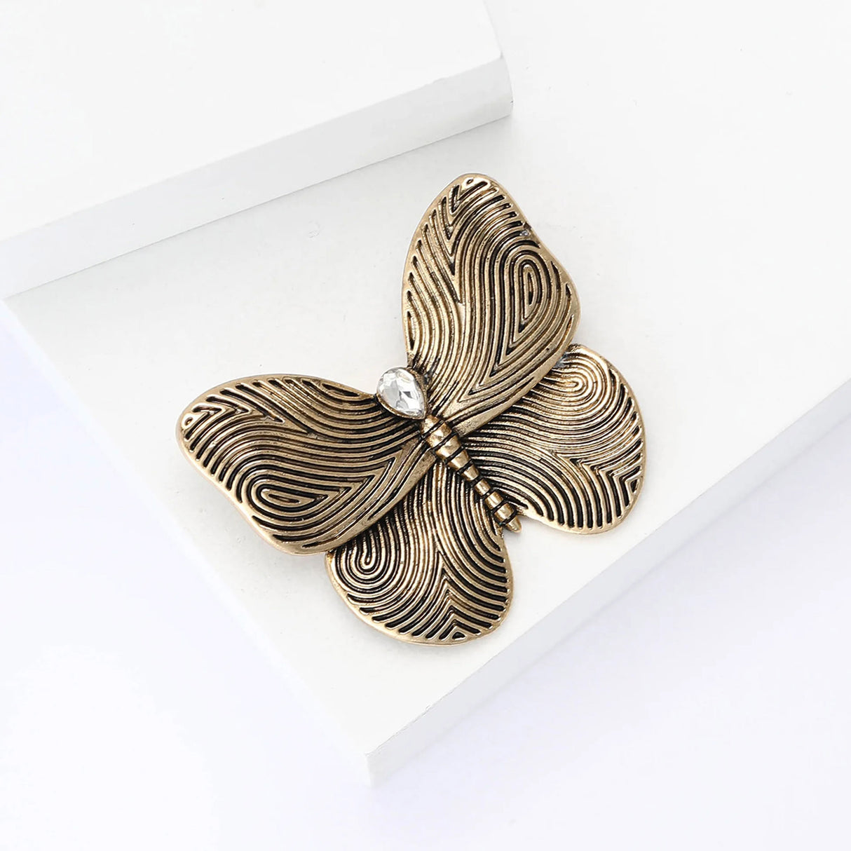 Butterfly brooch with gold-tone metal finish, textured swirl wings, and clear teardrop stone, flat lay on white studio surface.