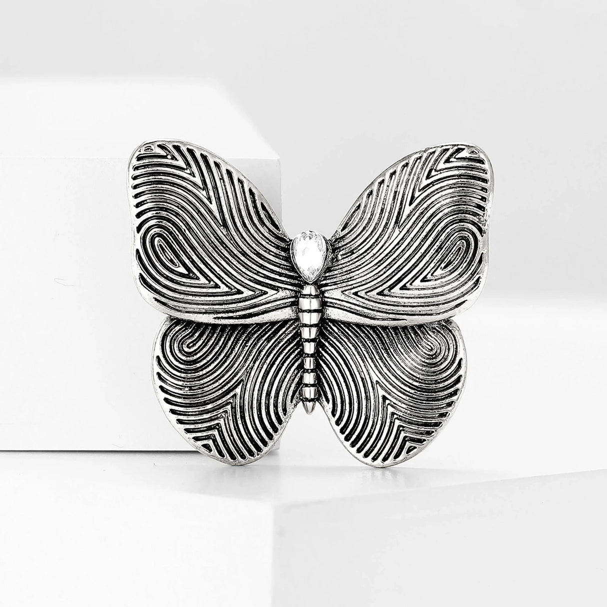 Butterfly brooch in silver-tone finish with etched swirl wings and clear teardrop stone, upright on white block.